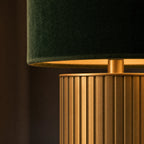 Detail of the Albany lamp — close-up of the brushed brass fluted column and the rolled bottom edge of the deep forest-green velvet drum shade, warm directional light catching the brass flutes and the soft pile of the velvet