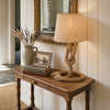 Aldeburgh hemp-rope and natural-linen drum table lamp on a slim aged-oak hallway console in an English countryside cottage, beside a stack of vintage cloth-bound novels, a tarnished-silver-rimmed gilt picture frame and a stoneware jug of dried lavender and bleached wheat, with a chalky tongue-and-groove panelled wall and antique gilt mirror behind