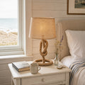 Aldeburgh hemp-rope and natural-linen drum table lamp on a chalky off-white painted bedside table in a Suffolk-coast weatherboarded fisherman's cottage guest bedroom, with linen bedding, an open paperback, a stoneware mug, a brass pocket-watch and a glass vase of wildflowers, beside a sash window onto a shingle beach and grey-blue North Sea