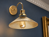 Belgravia detail close-up showing polished-brass S-curve gooseneck arm, brass collar with manufacturer label and small rivet detail, and wide amber-tinted fluted-glass coolie-hat shade with vertical flutes and Edison filament glowing inside, mounted on a Hague Blue plaster wall with Georgian dentil cornice
