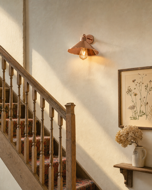 Cheltenham polished rose-copper short-arm wall light with flared coolie-hat shade and Edison filament bulb mounted on warm cream limewashed plaster wall midway up an oak Cotswold cottage staircase, with worn turned-spindle balustrade, Persian wool runner, framed botanical print and dried hydrangea in an ironstone jug