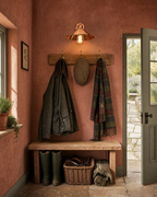 Cheltenham polished rose-copper cottage wall light mounted above an oak coat-hook rail with brass hooks holding a waxed Barbour jacket, tweed flat cap and tartan wool scarf, in a country cottage back-door boot room with terracotta clay-pink limewash plaster walls, stripped pine bench with green wellington boots, wicker basket of dog leads, and panelled door open onto stone path and herb garden