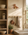 Cheltenham polished rose-copper cottage wall light mounted above a cream-painted Shaker peg rail with copper saucepan and striped tea towel, in an English country kitchen pantry corner with hand-plastered creamy-off-white limewash, deep stone Belfast farmhouse sink with brass kitchen tap, stripped pine open shelving with ironstone mixing bowls, sourdough starter jar and vintage glass spice jars, terracotta pot of fresh rosemary, golden-hour sunlight from a sash window