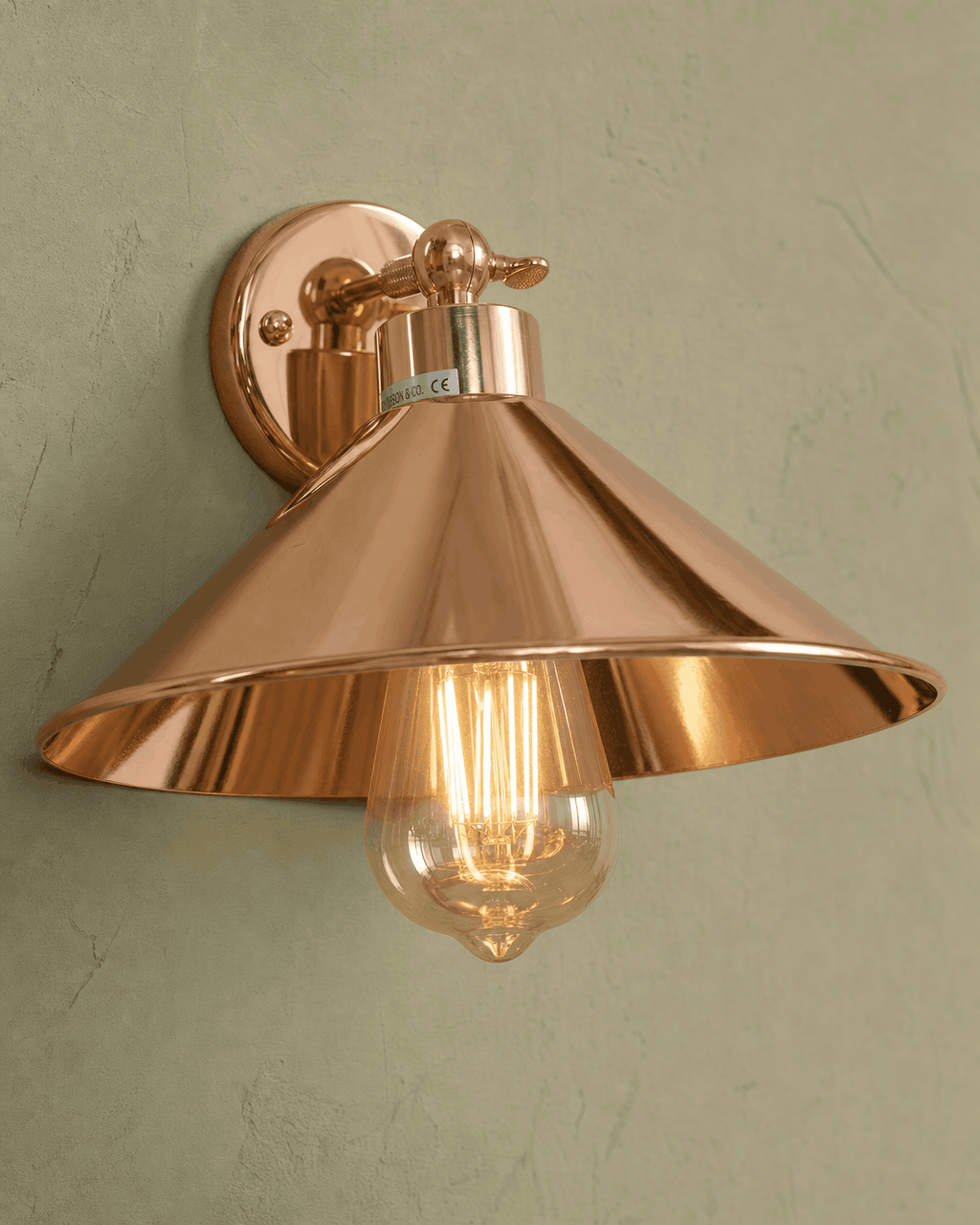Cheltenham detail close-up showing highly polished mirror-bright rose-copper electroplated finish across the small disc wall plate, short straight stem with knurled adjustment knob/collar at the joint, flared coolie-hat shade with bell-mouth rim and polished interior, and clear amber-tinted ST64 Edison filament bulb hanging straight down through the centre, mounted on hand-plastered sage-green limewash