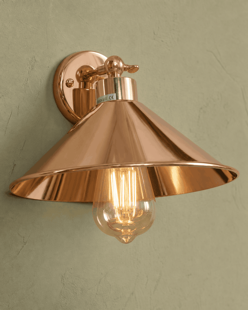 Cheltenham detail close-up showing highly polished mirror-bright rose-copper electroplated finish across the small disc wall plate, short straight stem with knurled adjustment knob/collar at the joint, flared coolie-hat shade with bell-mouth rim and polished interior, and clear amber-tinted ST64 Edison filament bulb hanging straight down through the centre, mounted on hand-plastered sage-green limewash