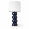 Cosmati bobbin ceramic table lamp on white background — stacked rounded ceramic forms in indigo high-gloss glaze beneath a crisp white drum shade