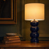 Cosmati bobbin ceramic table lamp in deep indigo glaze on a console table in a panelled English drawing room at dusk, with a crisp white drum shade pooling warm light onto framed art and bound books