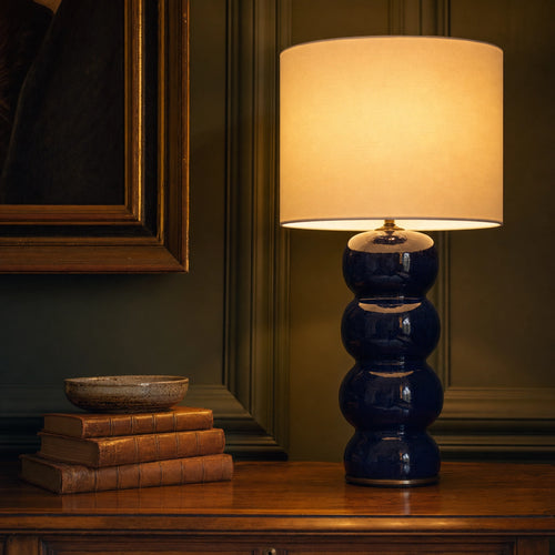 Cosmati bobbin ceramic table lamp in deep indigo glaze on a console table in a panelled English drawing room at dusk, with a crisp white drum shade pooling warm light onto framed art and bound books