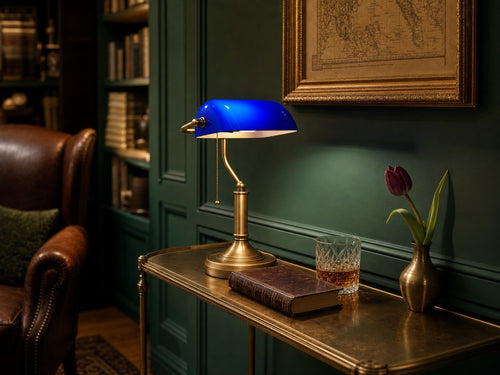 Lincoln Blue cobalt-glass banker's library table lamp on an antique brass console table beside a deep forest-green panelled wall, with an antique gilt-framed map, a leather-bound book, a cut-crystal whisky tumbler and a single tulip in a brass vessel