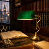 Lincoln Green emerald-glass banker's library table lamp on a leather-topped walnut writing desk in a panelled barristers' library, beside an open hand-written ledger, crystal inkwell and fountain pen, with leather-bound legal volumes and a sash window behind