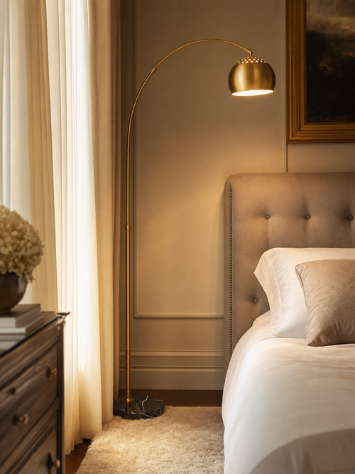 Mayfair brass arc floor lamp beside an elegant tufted bed with warm ambient lighting