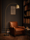 Mayfair brass arc floor lamp arching over a cognac leather reading chair in a dark library