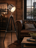Pinewood industrial searchlight tripod floor lamp casting warm light beside a leather Chesterfield sofa in a converted warehouse living room with exposed brick