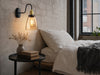 Whitechapel amber-glass gooseneck wall sconce mounted on a chalky off-white limewashed brick wall beside a low platform bed in a Victorian-warehouse-conversion bedroom